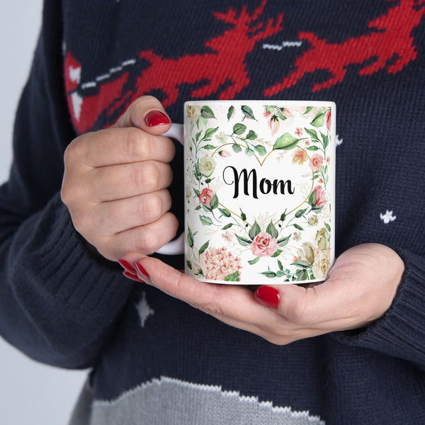 Person holding a mug with floral design and 'Mom' text, wearing a dark sweater with red deer pattern.