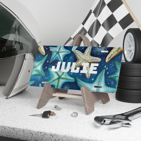License plate with starfish design and 'JULIE' text, placed on a stand with car accessories in the background.