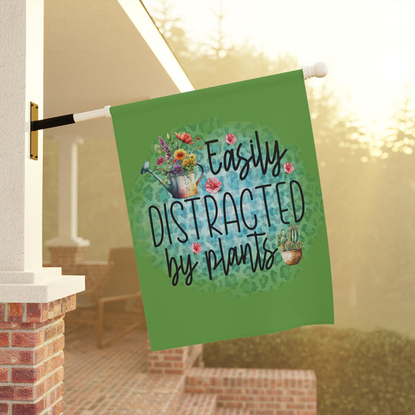Decorative garden flag with 'Easily Distracted by Plants' text and plant illustrations on a green background, hanging outdoors.