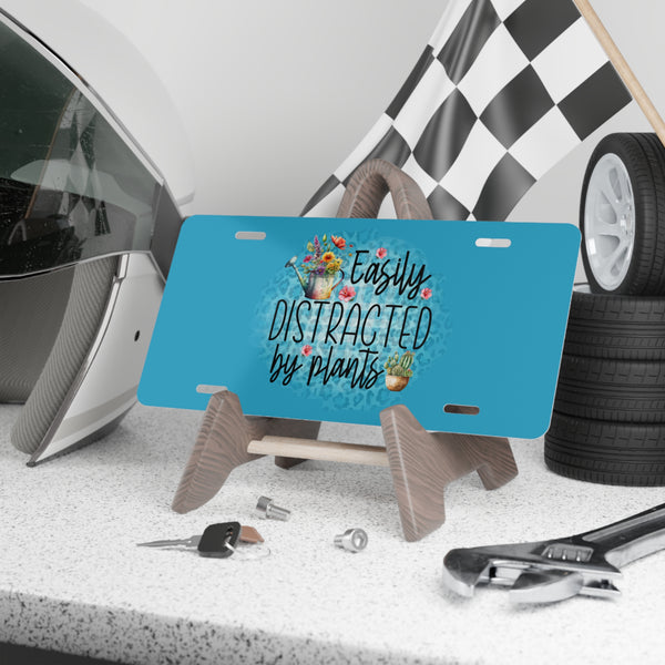 Blue license plate with 'Easily Distracted by Plants' text and plant illustrations on a stand, with a helmet, tools, and checkered flag in the background.