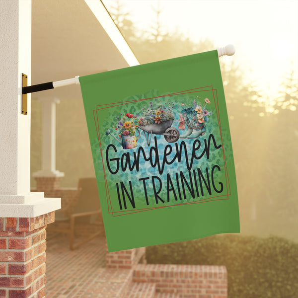 Decorative garden flag with 'Gardener in Training' text and gardening-themed design, hanging outside a house.