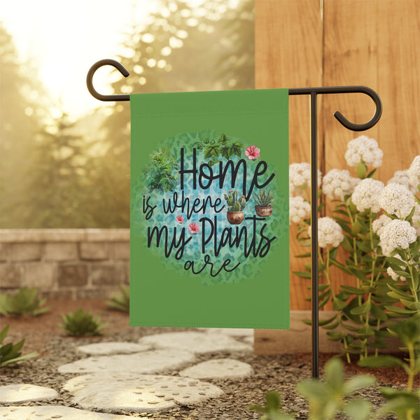 Garden flag with 'Home is where my plants are' text and plant illustrations on a stand with flowers and plants in the background.