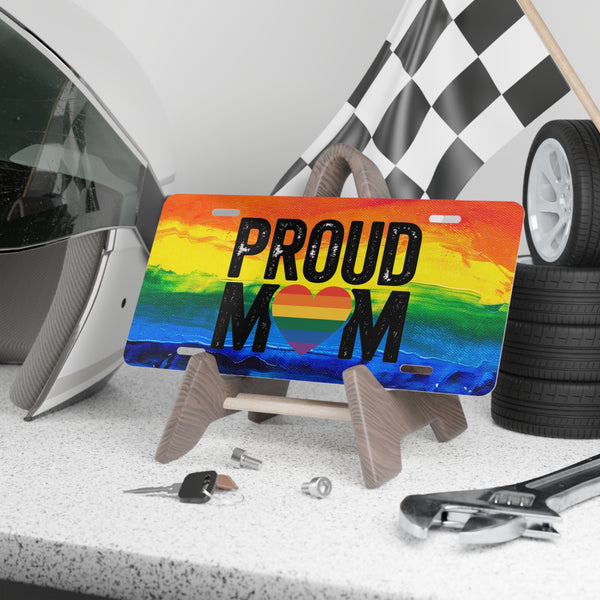 License plate with 'PROUD MOM' text on a rainbow background, placed on a stand with tools and car parts in the background.