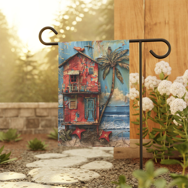 Decorative garden flag with a red house and palm tree design, displayed outdoors.