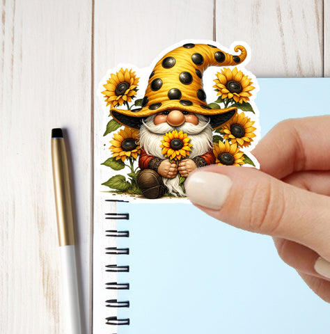 Sticker of a gnome with sunflowers held by a hand next to a pen on a notebook