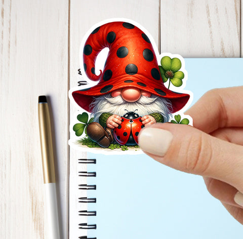 Sticker of a gnome with a red hat and ladybug held by a hand, on a notebook with a pen.